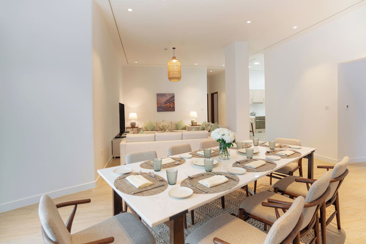 A dining area with a long table set for six, surrounded by cushioned chairs. A chandelier hangs above, and a sofa is visible in the background.