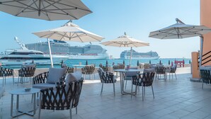 An outdoor seating area with tables and chairs, large umbrellas, and a view of cruise ships in the background. - Mina Hotel and Residences By the Torch (Doha)