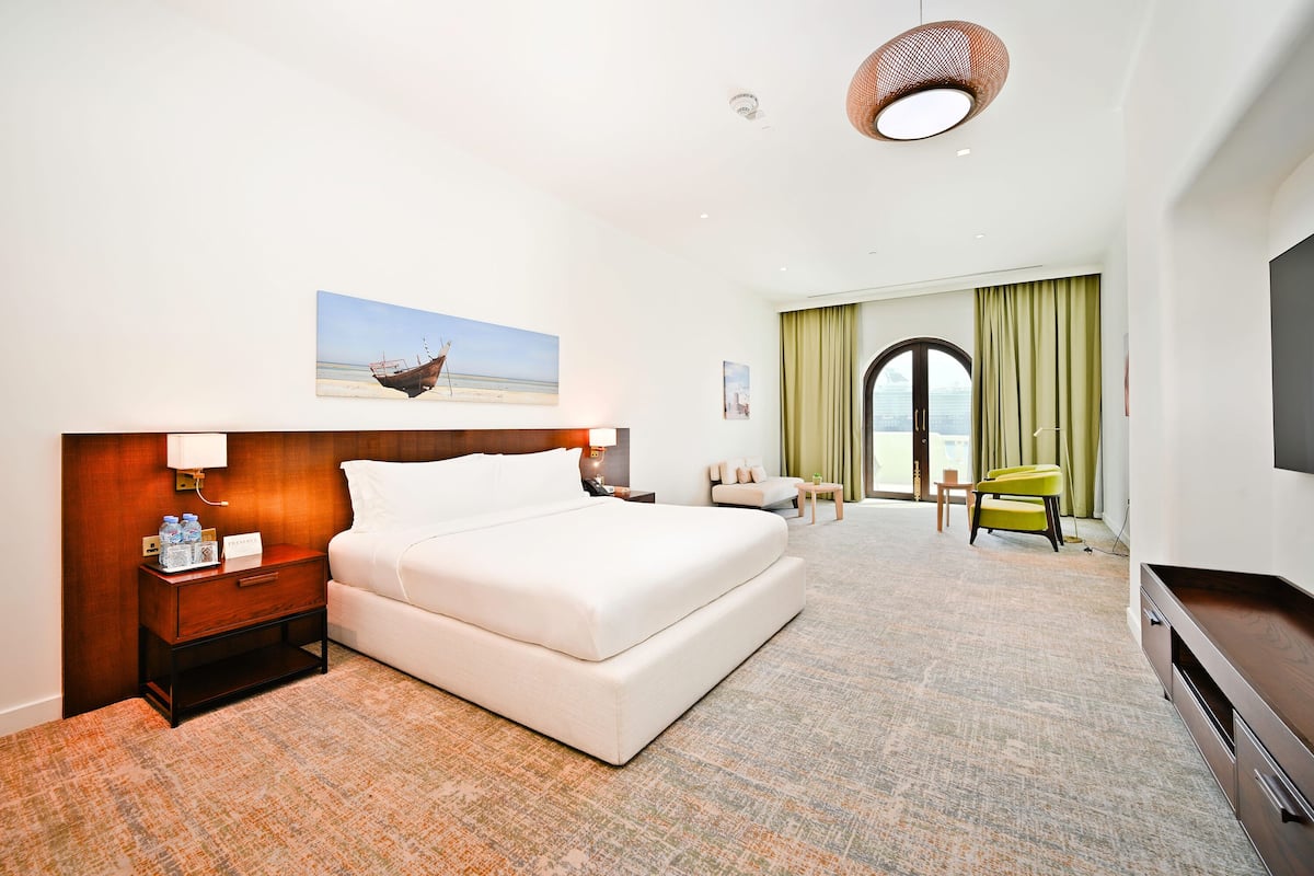 A hotel room with a bed, a nightstand, a TV, a painting, and a view of the sea.