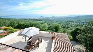 A terrace with a table and chairs, an umbrella, and a view of a forested landscape with a body of water in the distance. - Villa Maggiorana 10 (Lisciano Niccone)