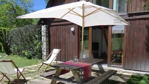 Un patio exterior con mesa y sillas de madera, un paraguas blanco y un muro de piedra al fondo.