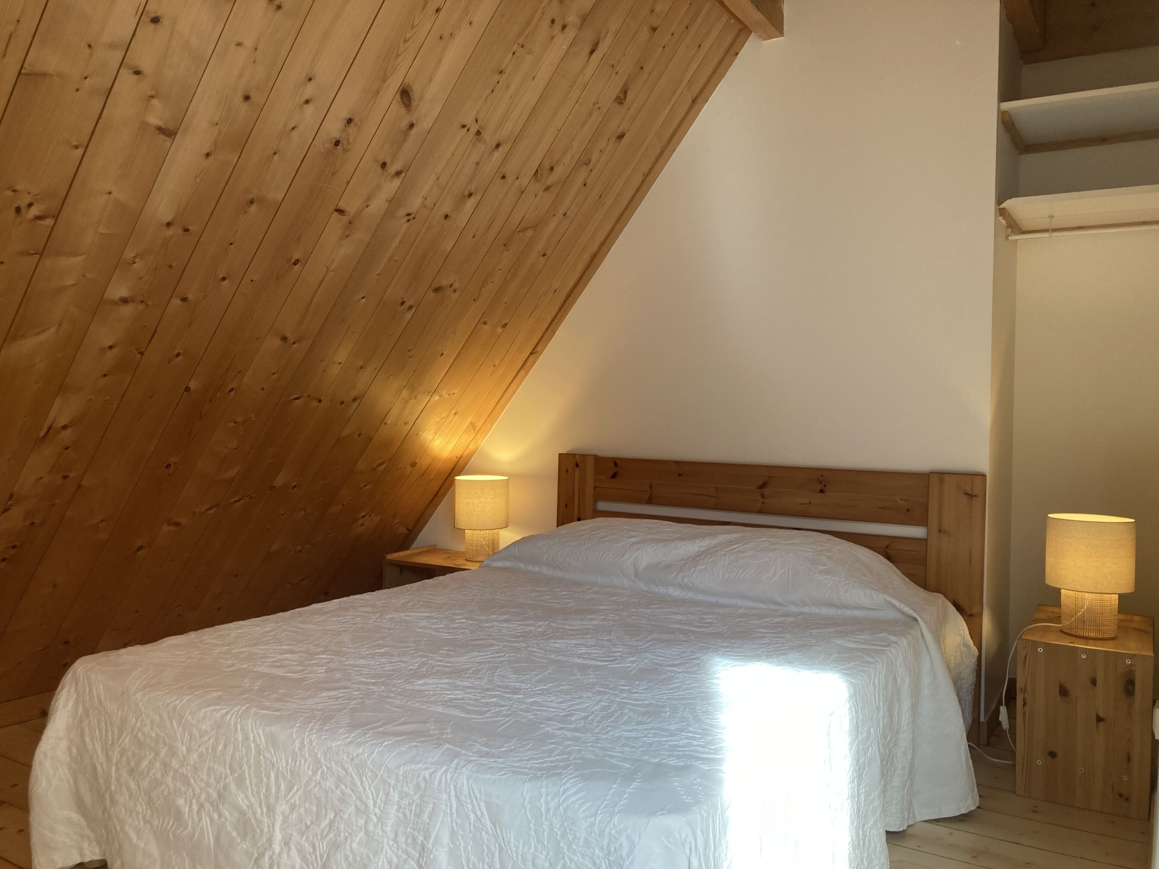 Een slaapkamer met een schuine plafond, houten hoofdbord, twee bedlampen en een bed met een witte dekbedovertrek.