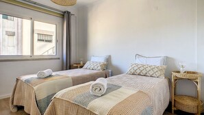A hotel room with two beds, a window, a bedside table with a lamp, and a wicker chair.