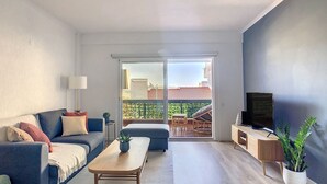 A modern living room with a sofa, coffee table, and TV. There is a sliding glass door leading to a balcony with outdoor furniture.