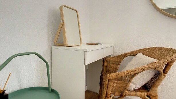 A white desk with a mirror and a notebook, a wicker chair with a cushion, and a green chair.