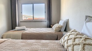 A double bed with a textured blanket, a bedside table with a lamp, and a window with a view of a hilly landscape.