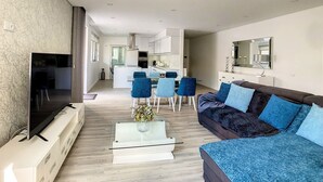 Un salon moderne avec un canapé, une table basse et un téléviseur. Au fond, on aperçoit une cuisine avec un coin repas.