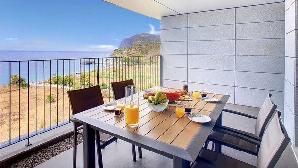 Un balcon avec une table préparée pour le petit-déjeuner, une vue sur l’océan et un paysage montagneux.