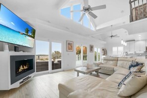 A spacious living room with a fireplace, a large flat-screen TV, a ceiling fan, and a view of the outdoors.