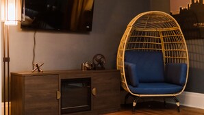 Une pièce avec un téléviseur fixé au mur, un fauteuil en osier avec des coussins bleus, une commode en bois et une lampe fixée au mur.