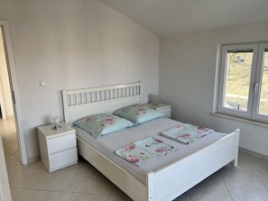 Une chambre avec un lit blanc, deux oreillers, une table de chevet et une commode, ainsi qu’une fenêtre donnant sur des arbres.
