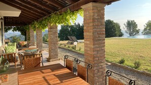 A covered outdoor seating area with brick pillars, a wooden deck, and a view of a grassy field and trees.