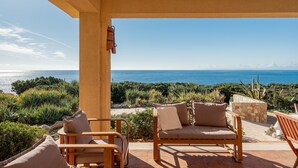 Un porche cubierto con dos sillas de madera y una mesa de piedra, con vistas a un paisaje costero con vegetación y el mar.