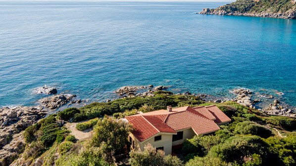 A coastal house with a red-tiled roof, surrounded by greenery and rocky terrain, overlooking a clear blue sea.