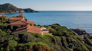 A coastal house with a red-tiled roof, surrounded by lush greenery and rocky terrain.