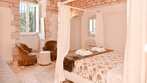 A bedroom with a four-poster bed, a sitting area with chairs, a radiator, and a window. - Masseria Profilo 10 (Monopoli)