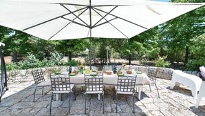An outdoor dining area with a table set for four under a large umbrella, surrounded by stone walls and greenery. - Masseria Profilo 10 (Monopoli)