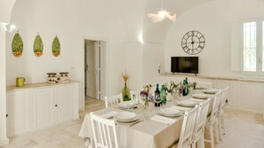 A dining area with a long table set for six, white chairs, a clock on the wall, and a TV above the table. - Masseria Profilo 10 (Monopoli)