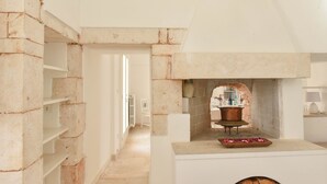 A stone-walled kitchen with a copper pot on a wooden stand, a white chair, and a small table with a flower arrangement. - Masseria Profilo 10 (Monopoli)