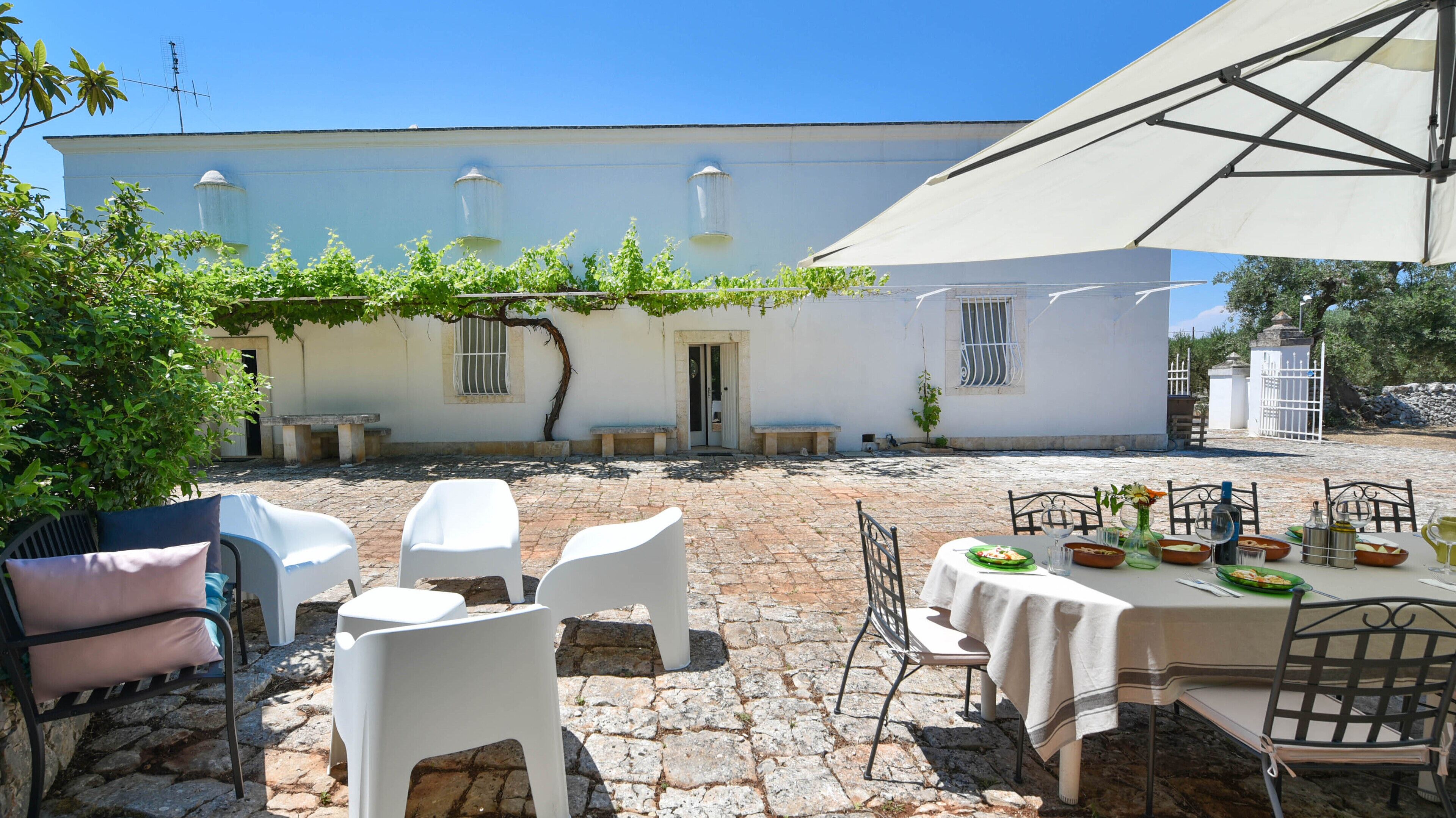A courtyard with stone flooring, white walls, and a white building with windows and a door. There are outdoor tables and chairs, and a pergola with vines.