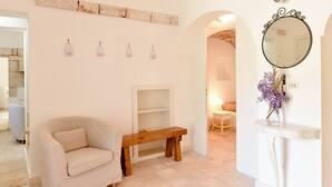 A room with a white armchair, a wooden bench, a mirror with a decorative frame, and a vase with purple flowers on a white console table. - Masseria Profilo 10 (Monopoli)