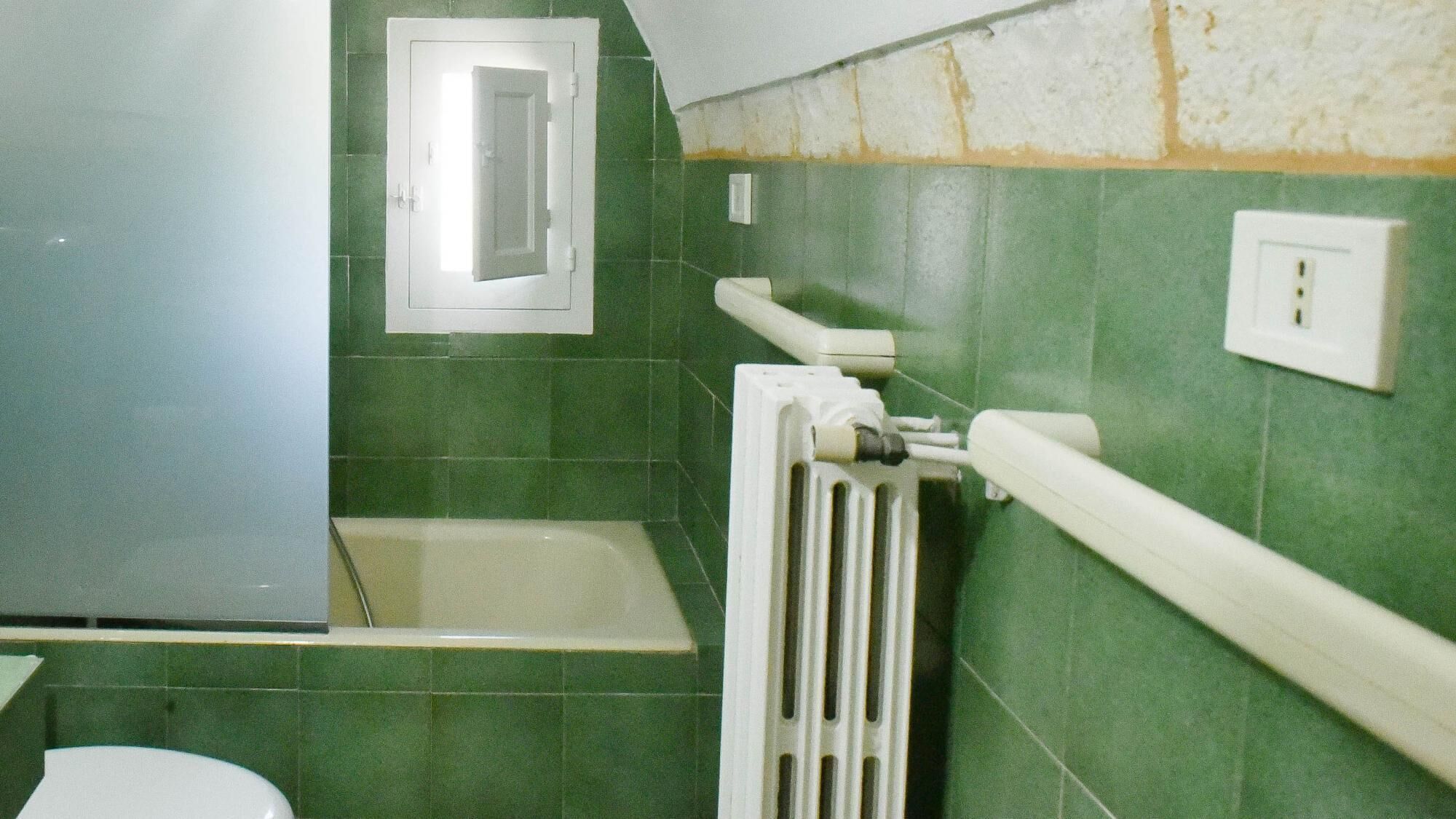 Un baño con azulejos verdes, un radiador blanco, un inodoro blanco y un ventanal pequeño.