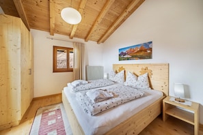A wooden bedroom with a bed, a nightstand, a radiator, a window with curtains, and a painting on the wall.