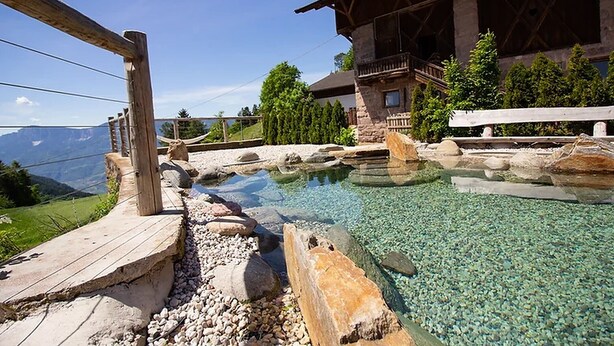 Una piscina nítida amb un pont de fusta, un edifici de pedra amb un balcó, i un paisatge de muntanyes al fons.