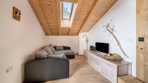 A loft-style room with a wooden ceiling, a large window, a sectional sofa, a TV on a wooden console, and a branch decoration.