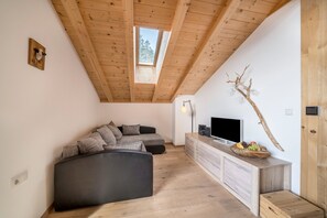 Ein Loft-Zimmer mit Holzbalkendecke, großem Fenster, Ecksofa, Fernseher auf einer Holzkonsole und einer Zweigdeko.