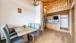 A dining area with a wooden table and blue upholstered chairs, a kitchen with wooden cabinets and a built-in stove, and a wooden staircase leading to an upper level. - Apartment "Eggerhof Fernblick" with Mountain View, Lake Access & Wi-Fi (Verano)