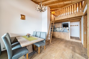 Un comedor con mesa de madera y sillas tapizadas en azul, una cocina con armarios de madera y estufa empotrada, y una escalera de madera que sube a un nivel superior.