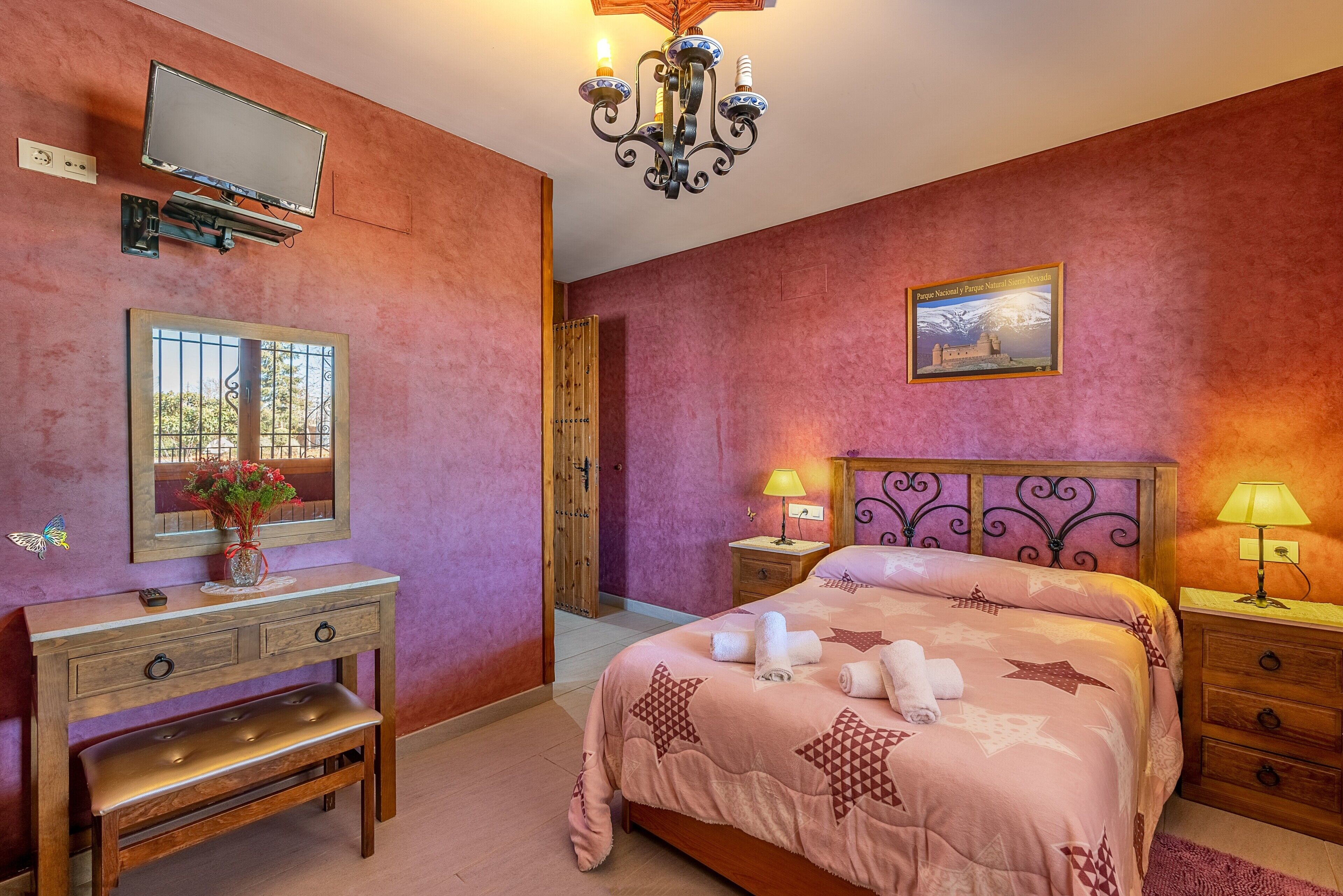 Un dormitorio con cama, mesitas de noche, tocador con espejo, banca, candelabro y un cuadro en la pared.