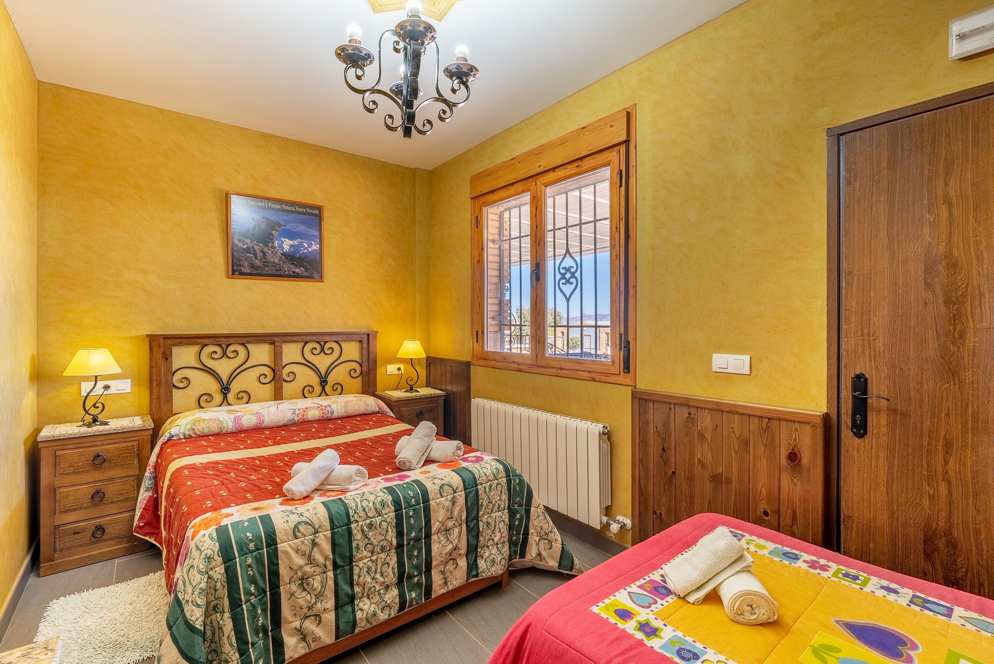 A bedroom with two beds, wooden headboards, a window with a view, a chandelier, and a radiator.