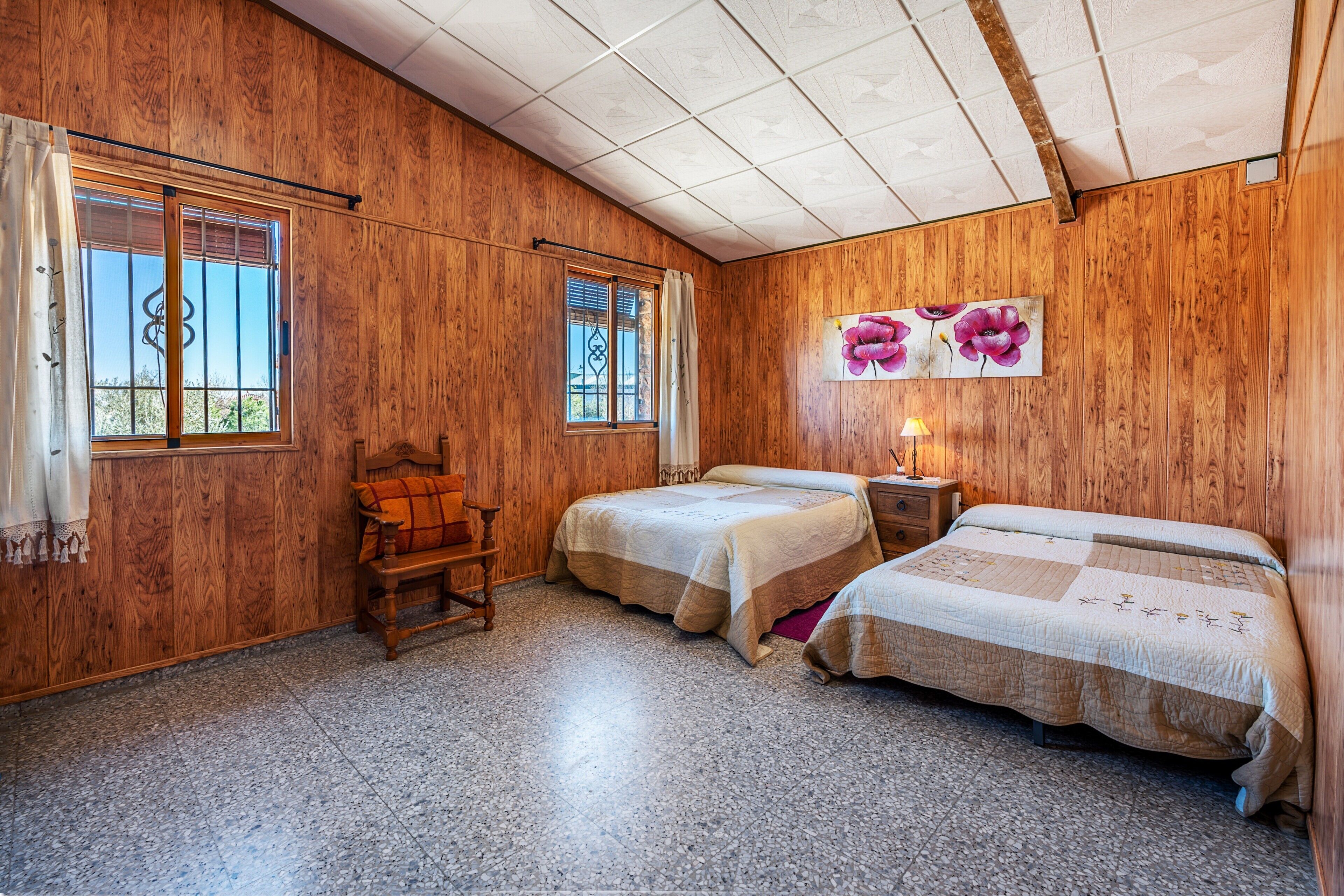 Una habitación con paredes de madera, dos camas, una silla y un ventanal con cortinas.