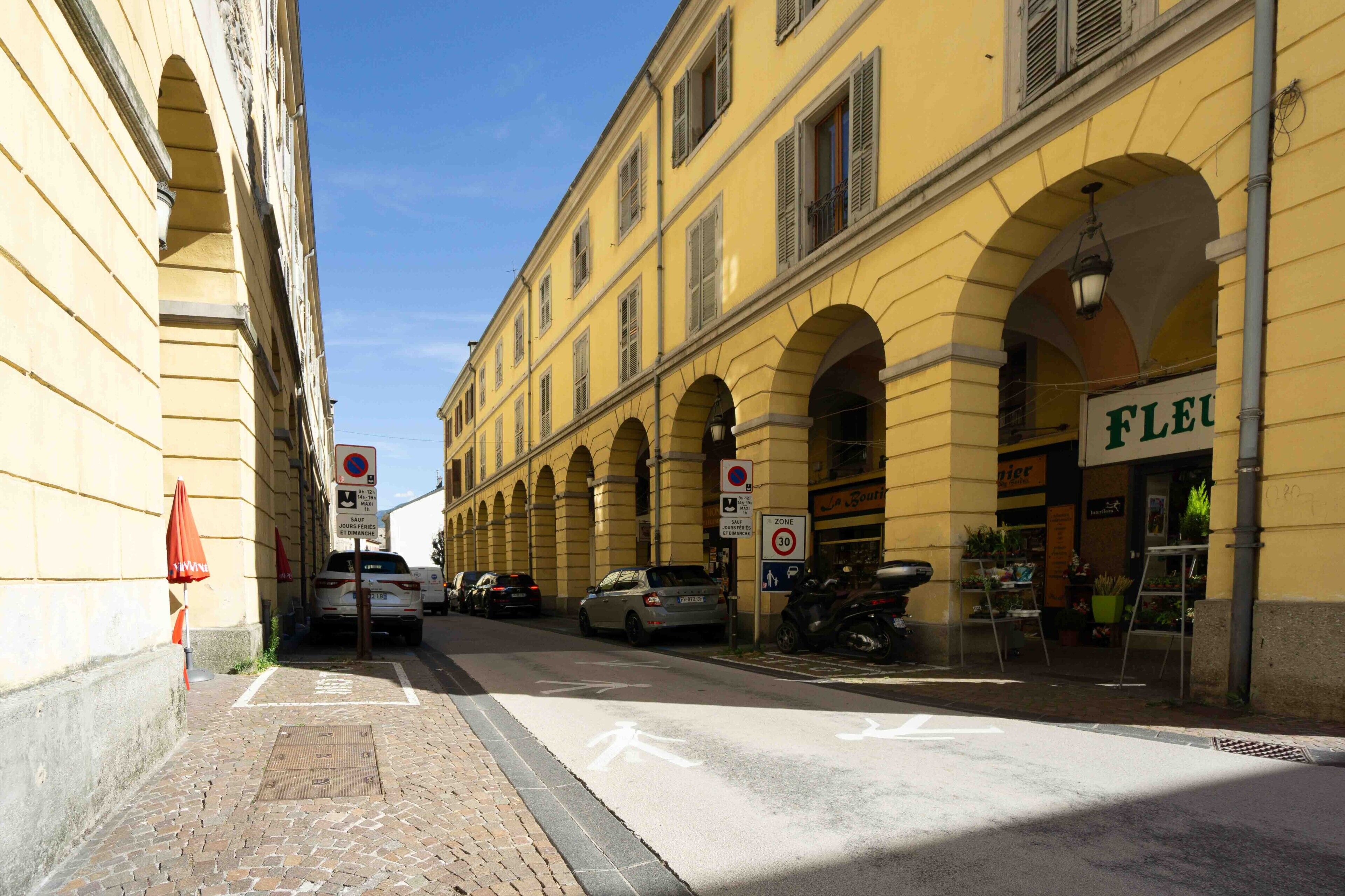 A street with yellow buildings, a 'FLEUR' shop, and a 'Zone 30' speed limit sign.