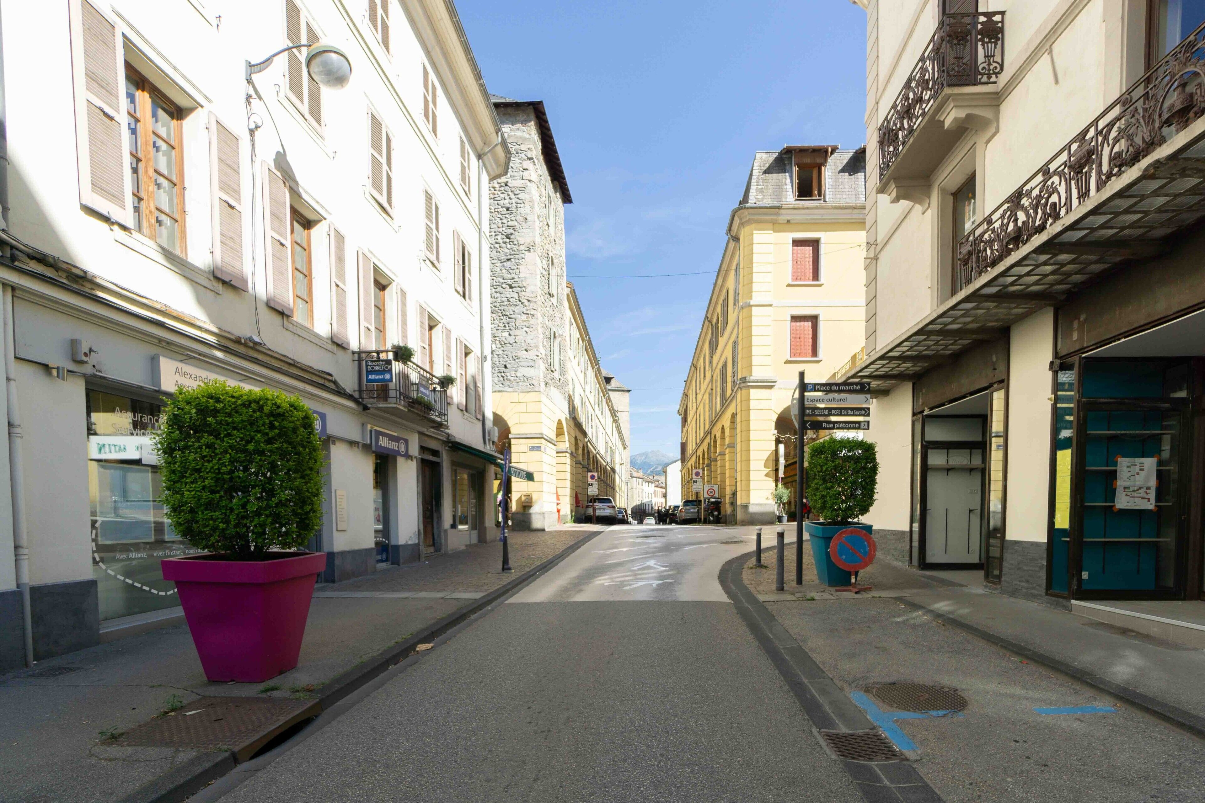 Une rue étroite, bordée de bâtiments des deux côtés, avec un grand pot de fleurs sur le côté gauche et un panneau indiquant l’interdiction de se garer sur le côté droit.