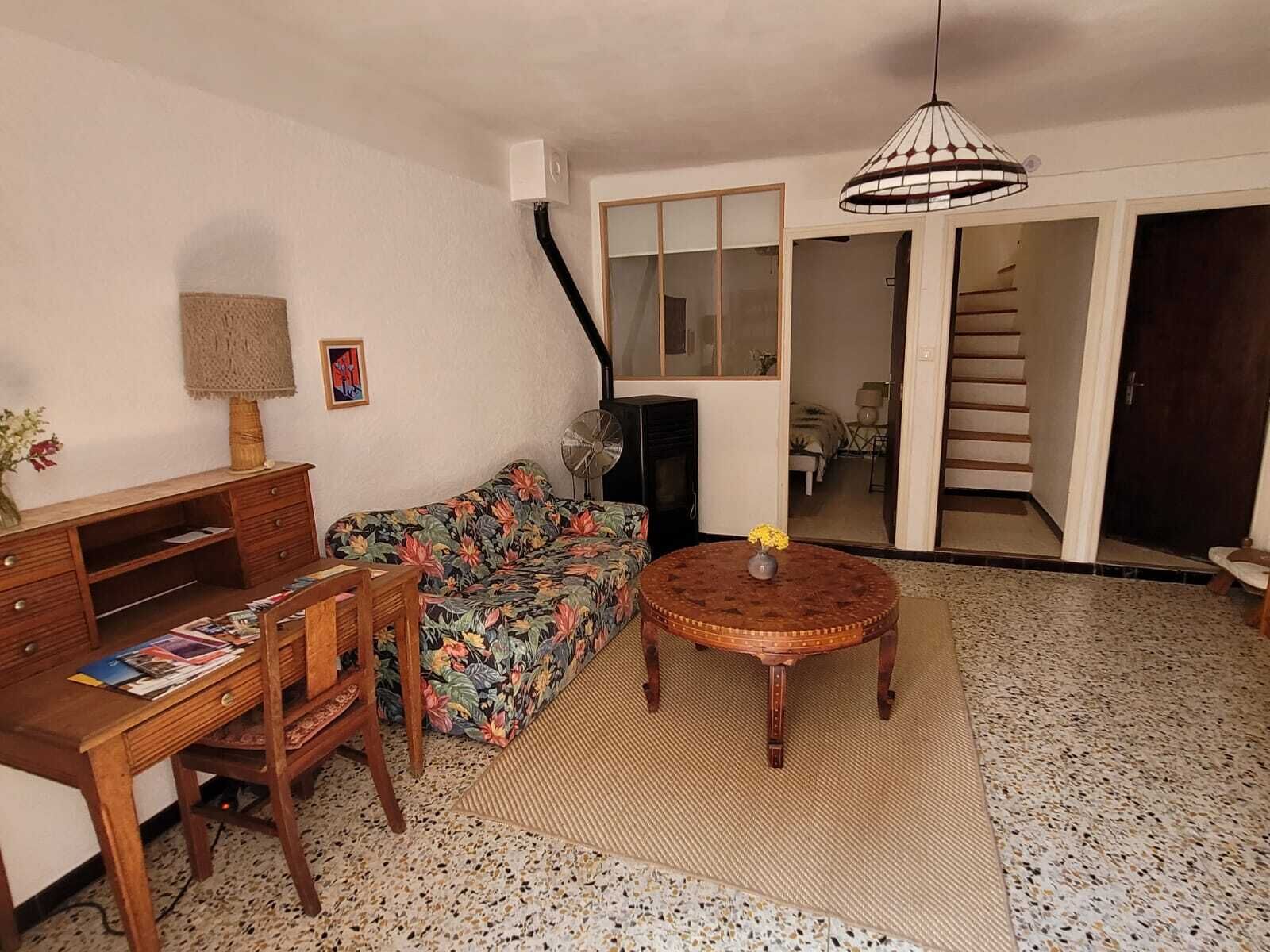 Un espace de vie compact comprenant un canapé, un bureau en bois, une petite table avec un vase, un ventilateur sur pied et un escalier menant à une autre pièce.