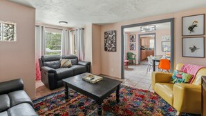 Una sala de estar con un sofá de cuero negro, un sofá amarillo, una mesa de centro y una alfombra colorida. Se ve una cocina al fondo con una mesa de comedor y sillas.