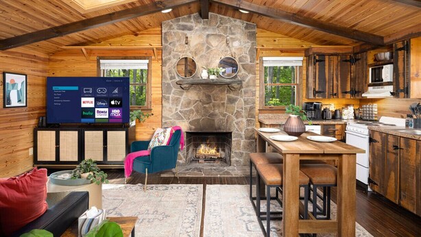 Un acogedor interior de cabaña con chimenea de piedra, vigas de techo de madera, un televisor y una zona de cocina.