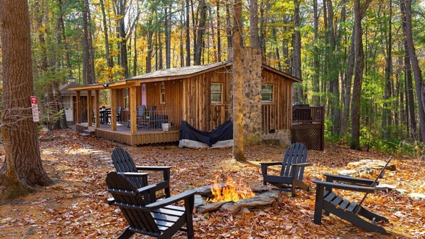 A cabin with a porch, a fire pit, and Adirondack chairs in a forest.