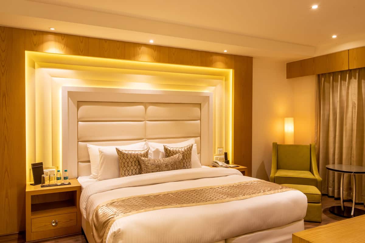 Habitació d'hotel amb un llit gran, taules de nit, un otomà verd, una taula rodona petita i una finestra amb cortines.