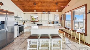 Une cuisine avec un plafond en bois, des armoires blanches, un grand îlot central avec des tabourets et une vue par les fenêtres.