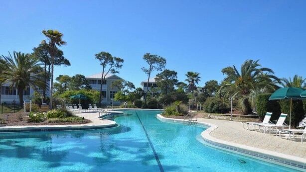 A clear swimming pool with lounge chairs and umbrellas, surrounded by palm trees and a building in the background. - Welcome to Good Day Sunshine! (St. George Island)