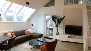 Una sala de estar moderna con sofá, mesa de centro y televisor. Se ve una escalera y un balcón a través de la puerta.