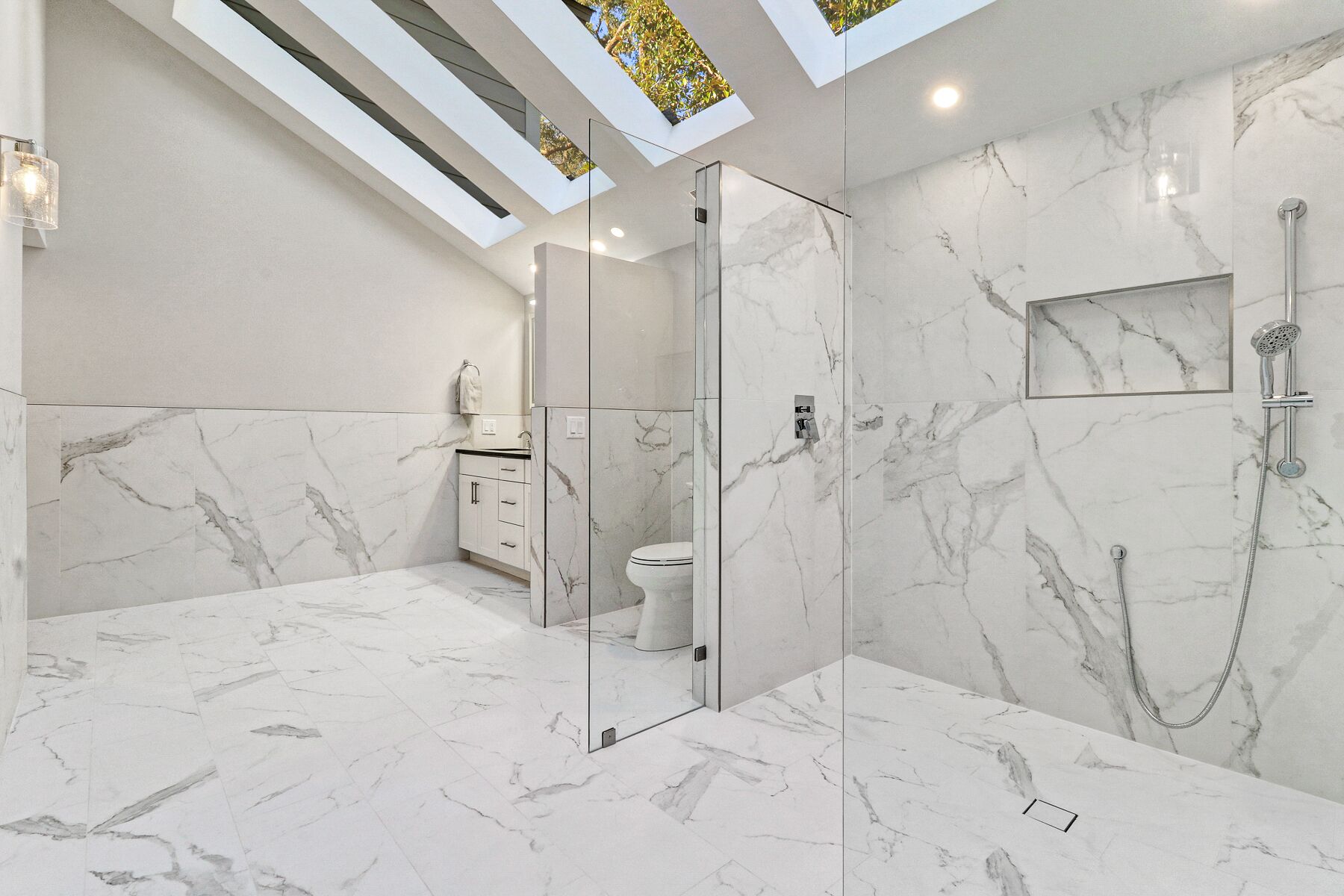 A modern bathroom with marble floors and walls, a glass-enclosed shower, and skylights.