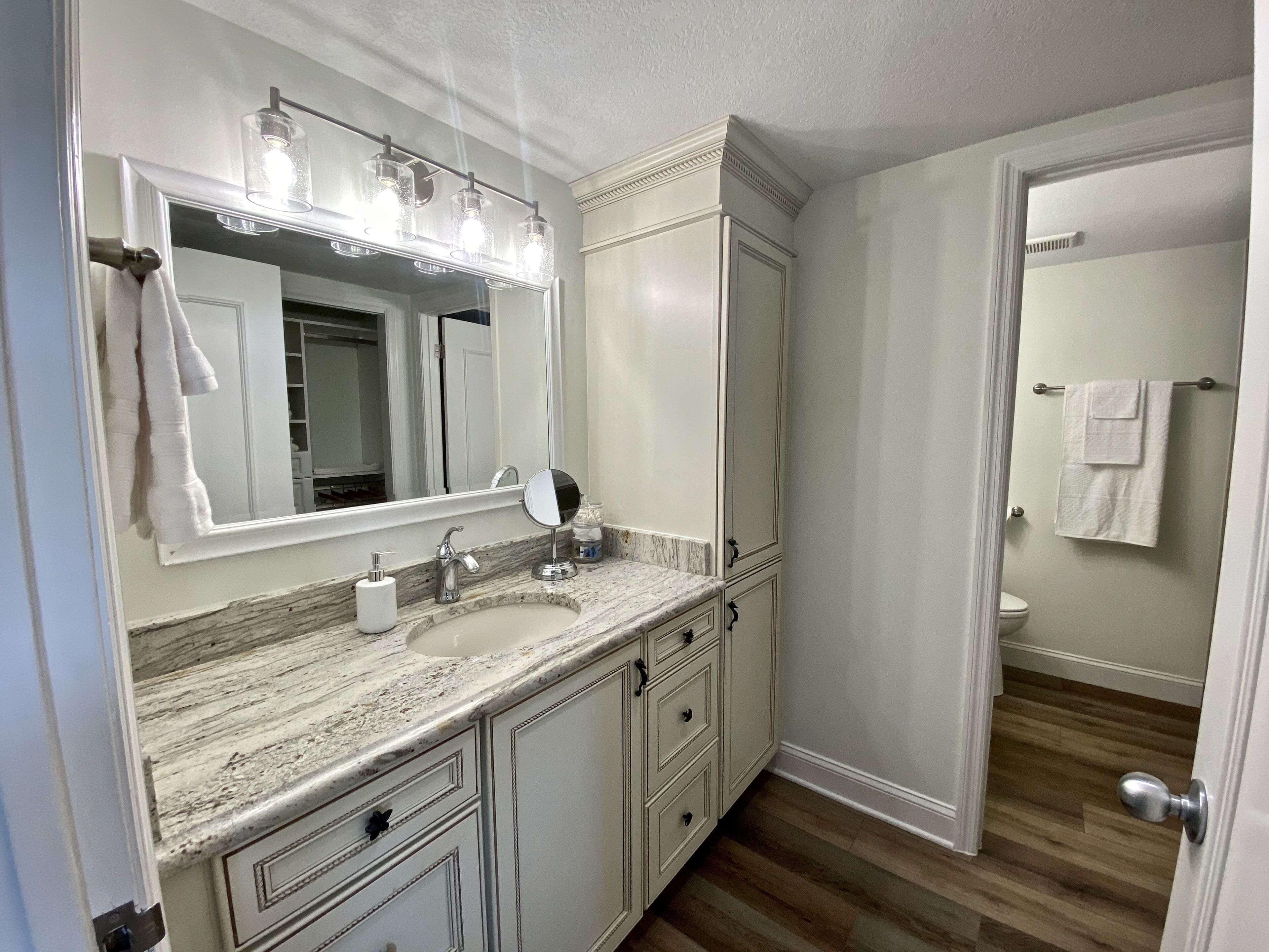 A bathroom with a marble sink, a large mirror with lights, a toilet, and a door leading to another room.