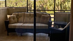 A room with a cushioned sofa and a small table, large windows with a view of palm trees and a body of water.