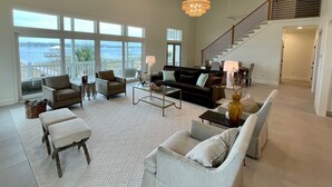 A spacious living room with a large window overlooking a body of water, a ceiling fan, and a staircase leading to an upper level.
