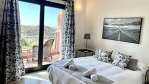A hotel room with a large bed, a view of the countryside, a painting on the wall, and a balcony with a chair. - Villa Over the 5th Green (San Bartolomé de Tirajana)